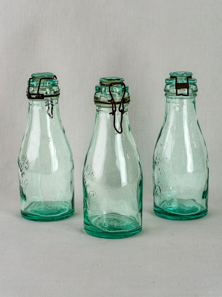 Set of three empty La Lorraine antique French aqua glass preserving bottles arranged on a neutral surface.