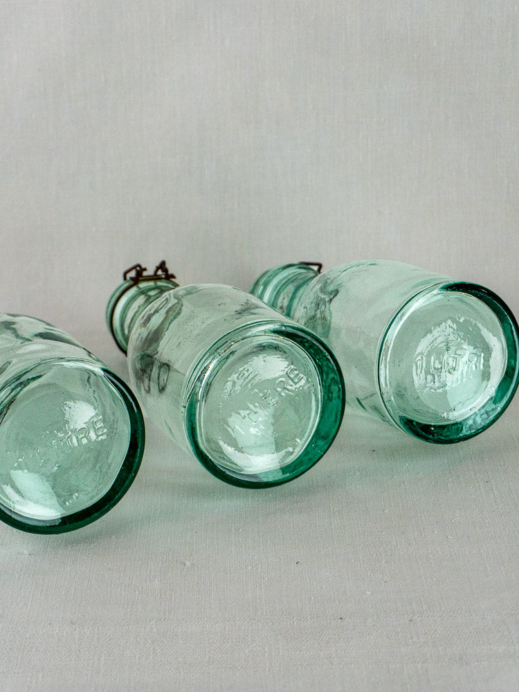 Three antique French aqua glass preserving bottles with embossed bases on a light surface.