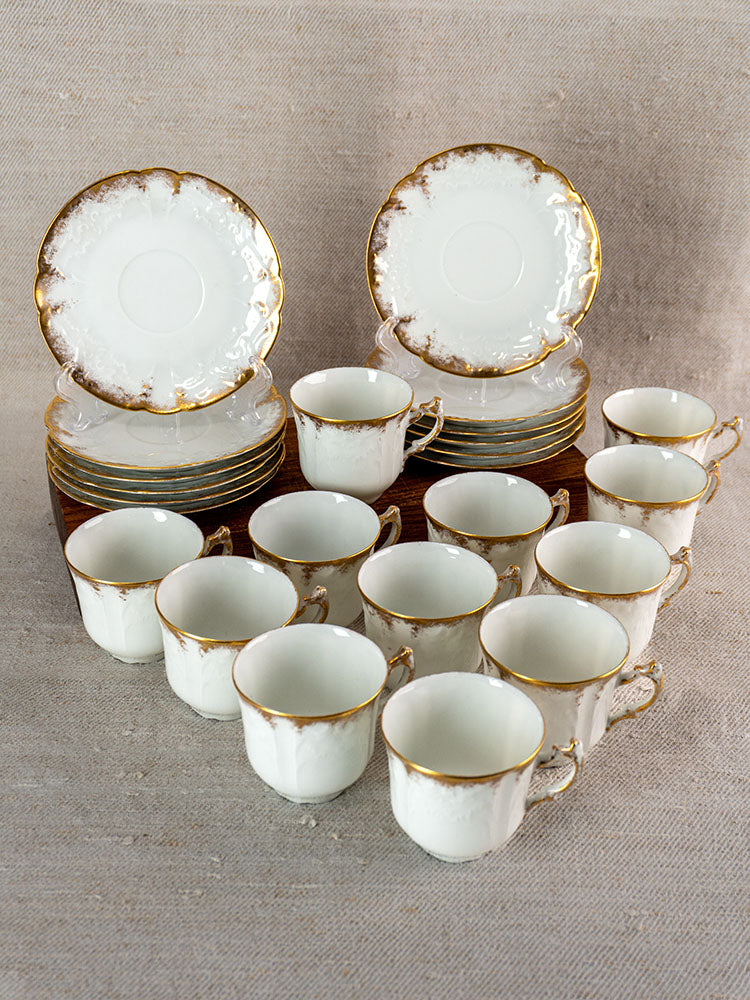 Set of twelve antique French Limoges demitasse cups with gold rims and matching saucers on beige fabric.