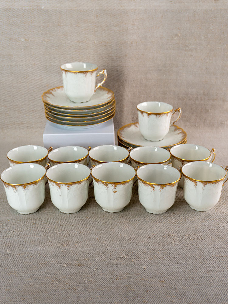 Antique French Limoges porcelain demitasse service with gold-trimmed cups, saucers on beige fabric.