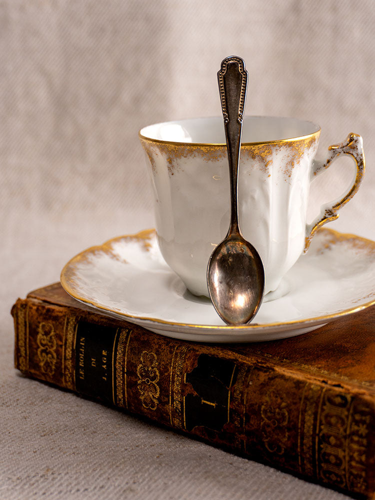 Antique French Limoges demitasse teacup and saucer with gold trim and tarnished spoon on ornate book.