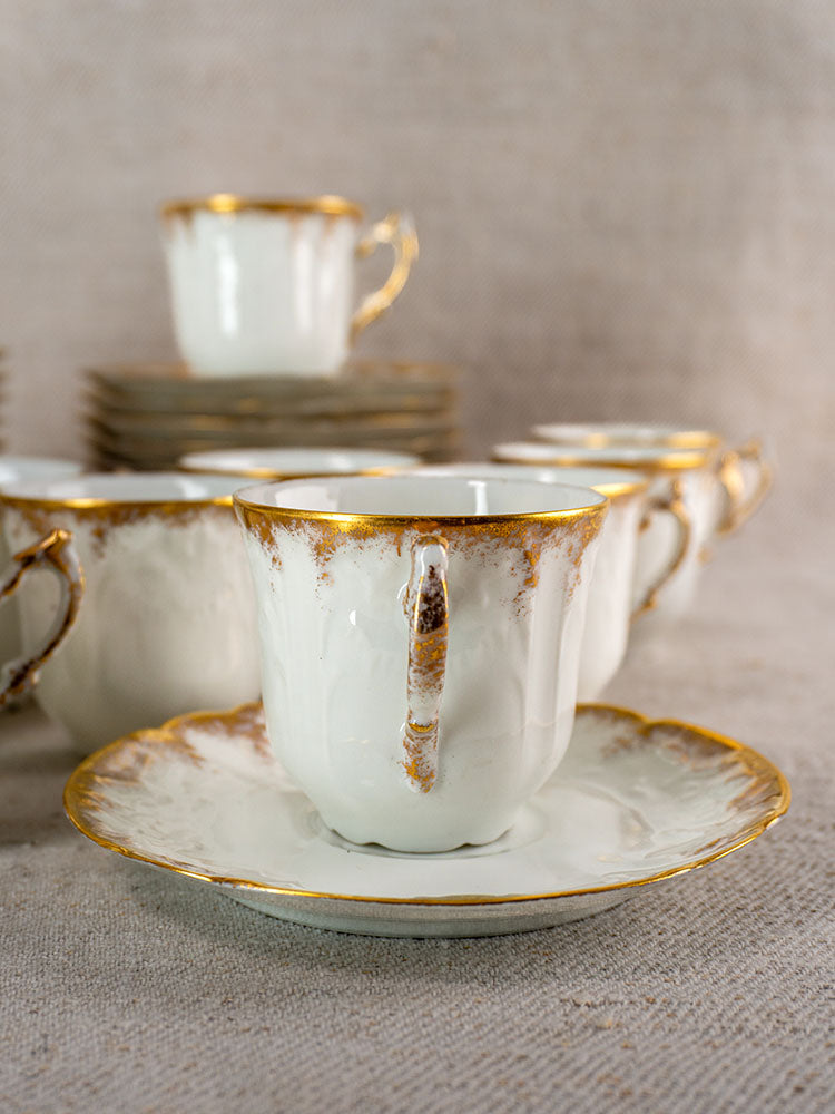 Antique French Limoges demitasse cups with gold trim on matching saucers, neutral fabric backdrop.