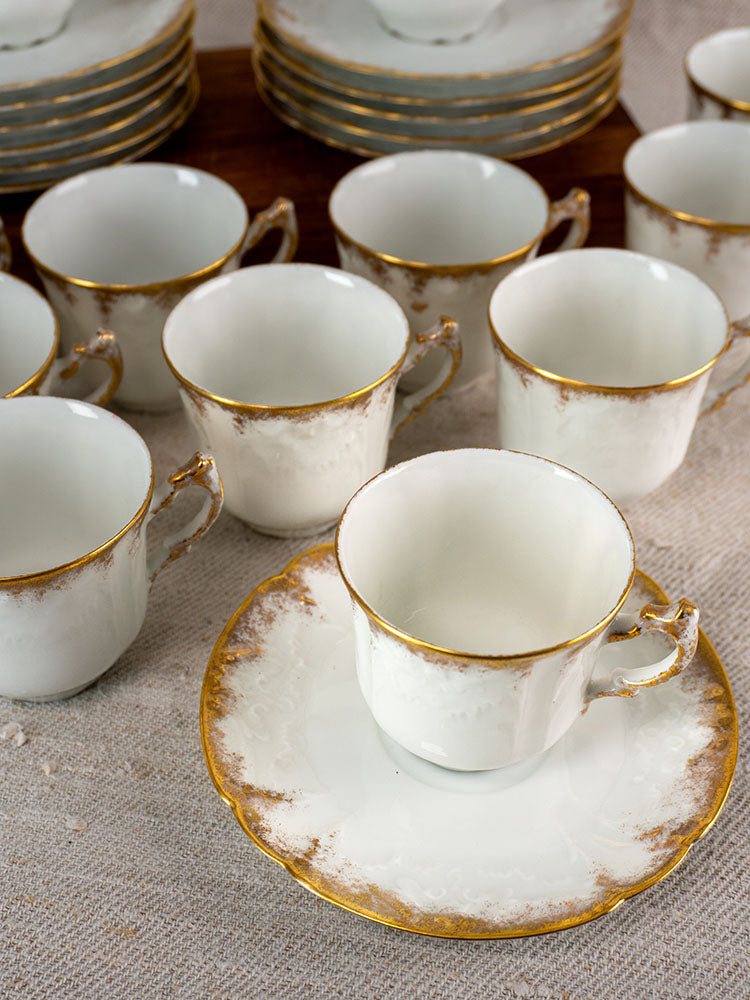 Antique French Limoges demitasse set for 12 by Lafarge & Cie, white porcelain with gold trim, on beige tablecloth.