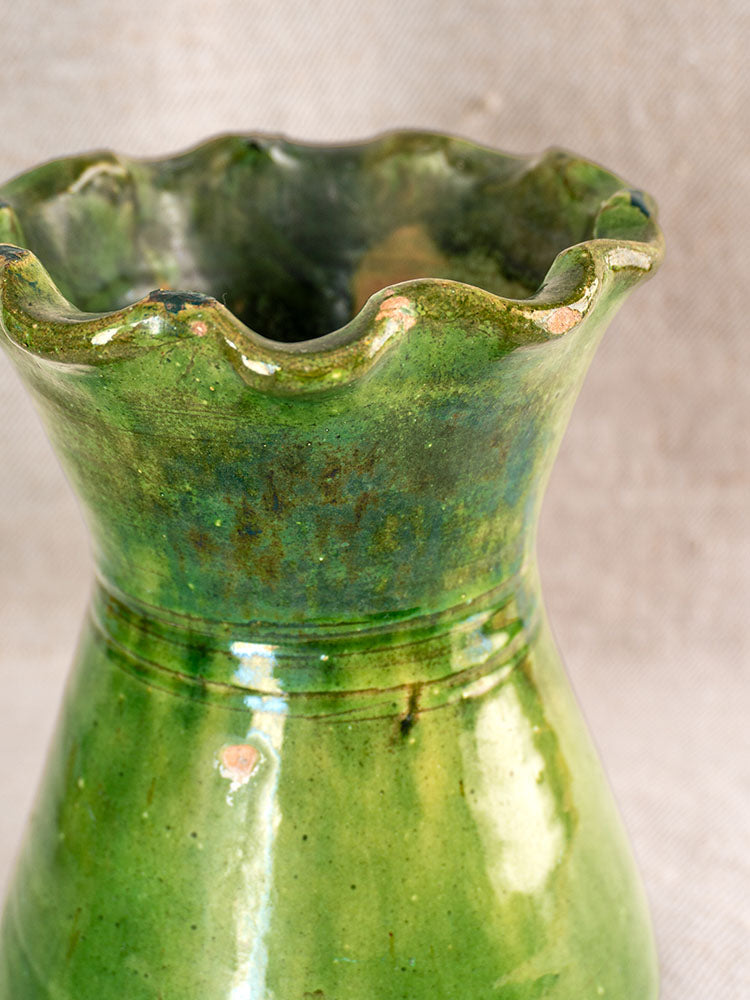 19th century French terracotta vase with green glaze, scalloped rim, worn finish; on neutral textured fabric background.