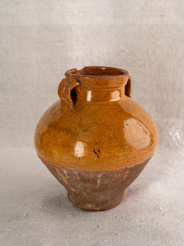 Antique French earthenware confit pot with small spout, dual handles, brown glaze, unglazed base on light fabric.