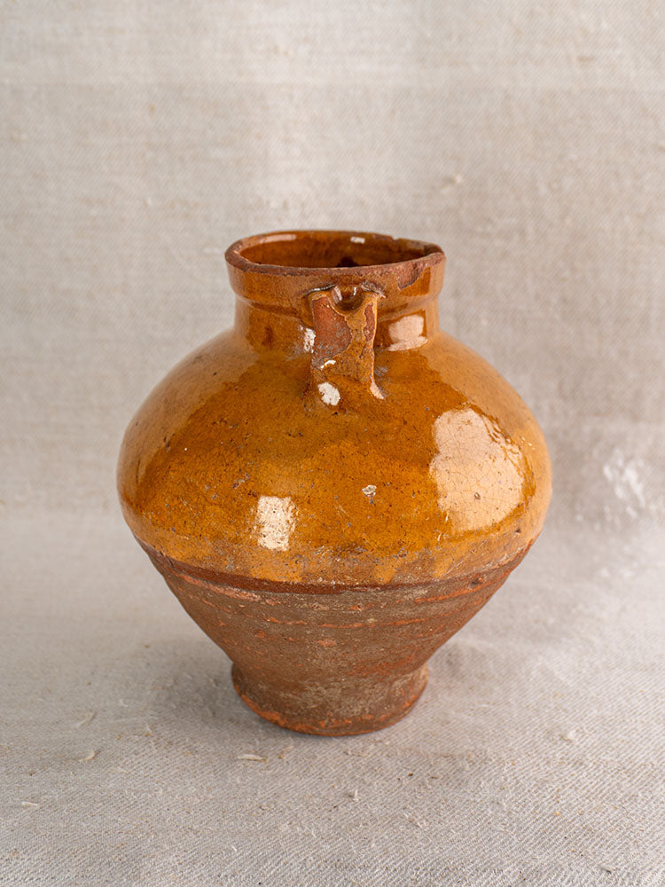 Antique French glazed earthenware olive oil jar with wide body, narrow neck, and small handles on beige textured fabric.