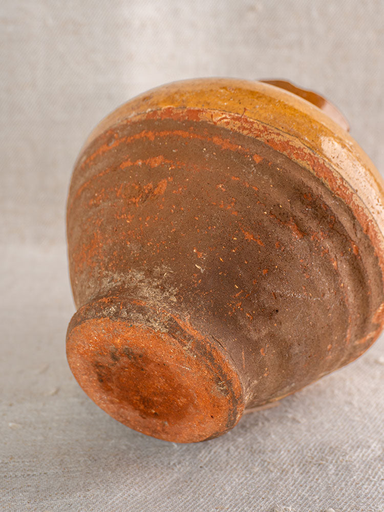 Antique French olive oil jar in glazed earthenware, reddish-brown base, resting on neutral fabric background.
