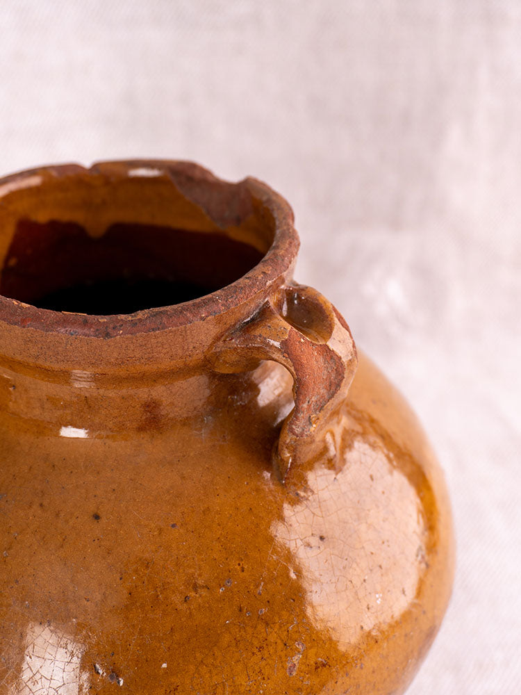 Antique French glazed earthenware olive oil jar with chipped rim and small handle on textured light fabric background.