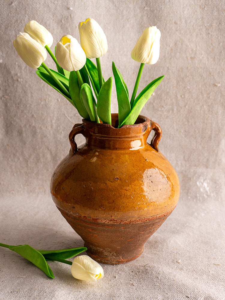 Antique French glazed earthenware olive oil jar with white tulips, displayed on textured beige fabric.