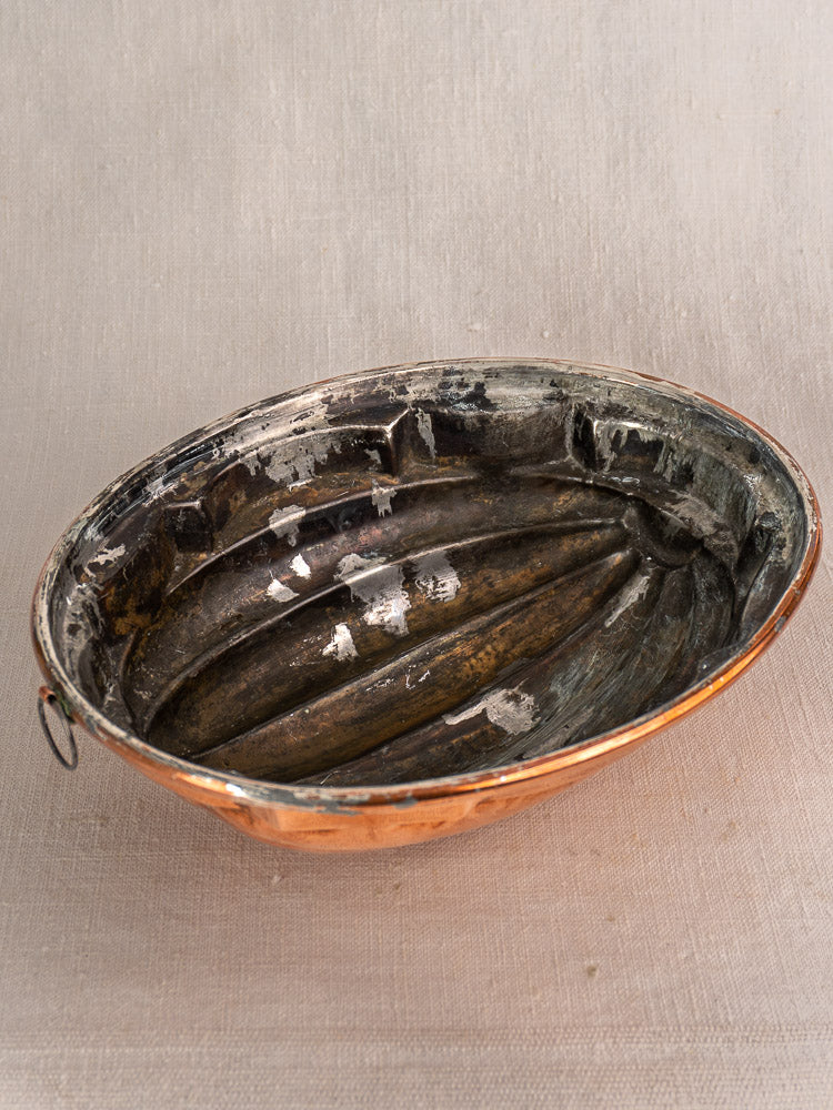 Antique French copper melon mold, oval tin-lined with worn interior and hanging loop, on textured beige surface.