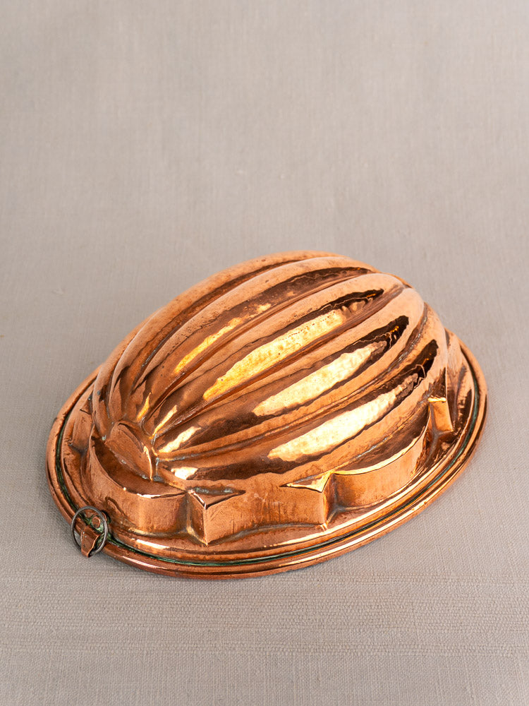 Antique French copper melon mold with ridged oval design and hanging loop on neutral fabric surface.