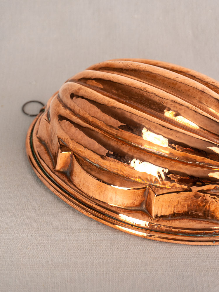 Antique French copper melon mold with ridged dome and hanging loop displayed on light beige fabric.