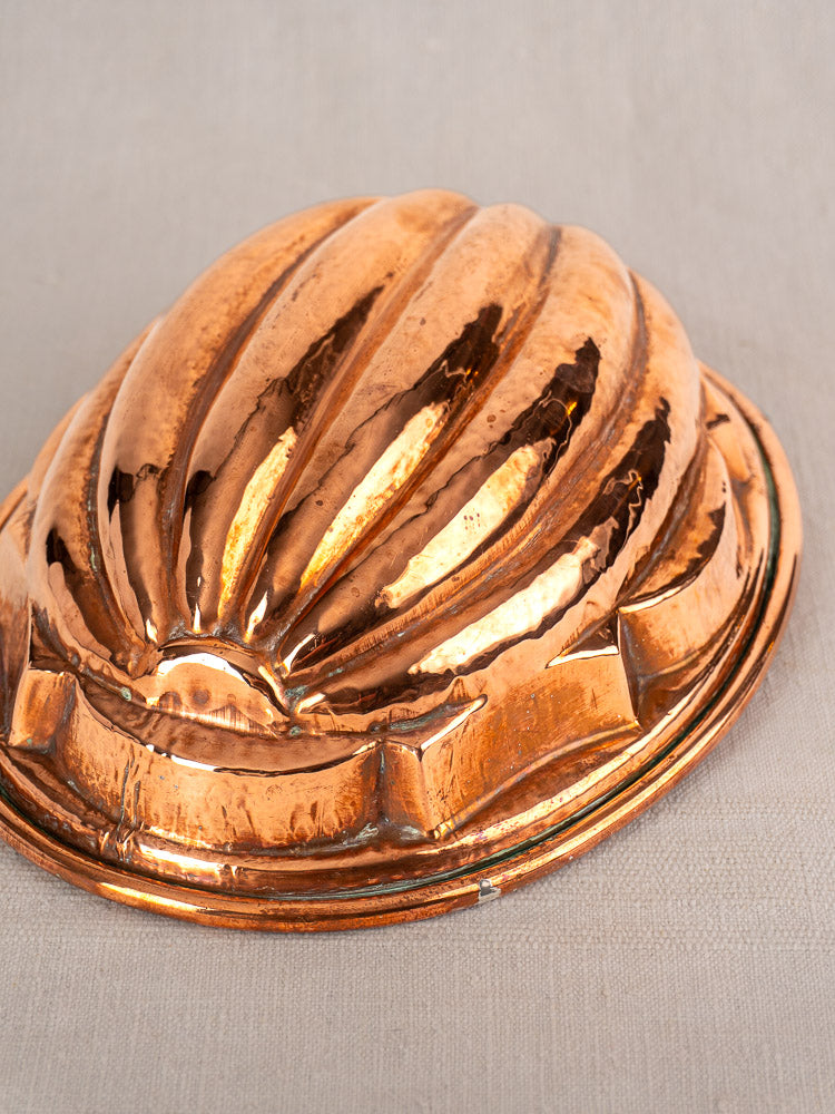 Antique French copper melon mold with ribbed shell pattern, tin-lined, on a pale beige fabric background.