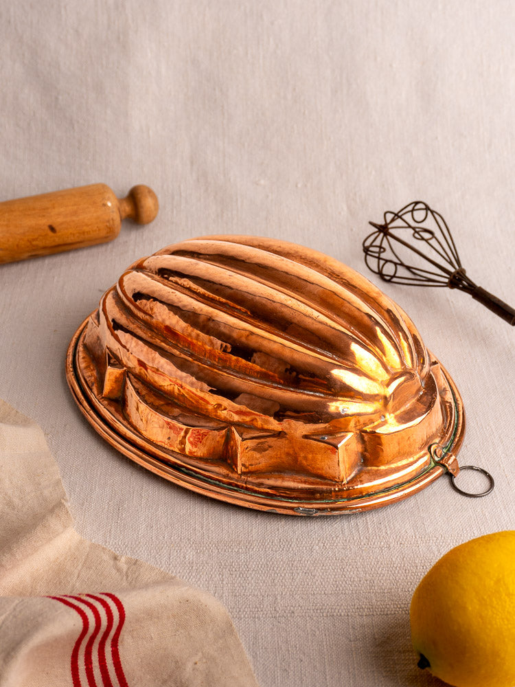 Antique French copper melon mold on light fabric, with rolling pin, whisk, striped cloth, and lemon for kitchen decor context.