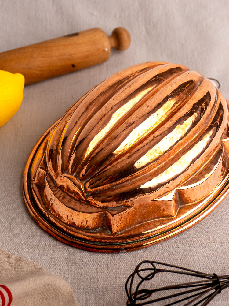 Antique French copper melon mold with tin lining, displayed on light fabric beside rolling pin, lemon, and whisk.
