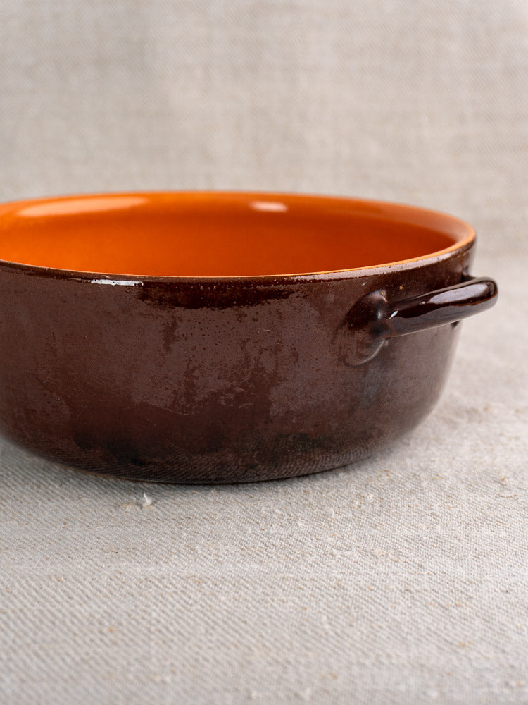 Antique French earthenware baking dish, glossy brown glaze, orange interior, side handle, on textured gray fabric. Mid 20th century.
