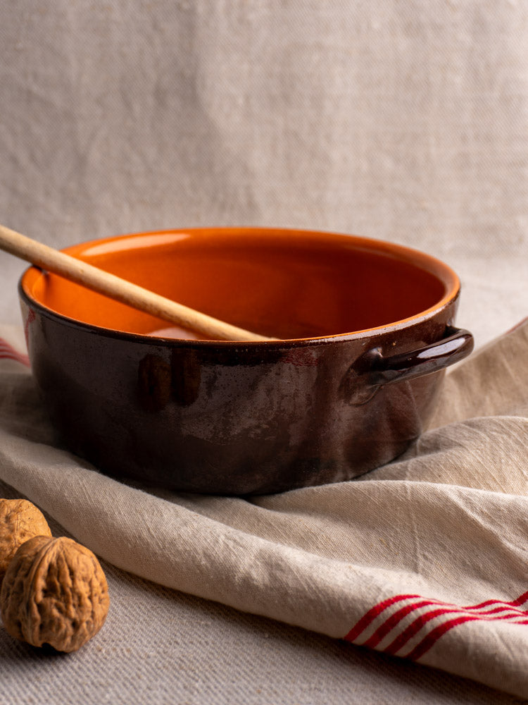 Antique French earthenware baking dish with brown and orange glaze on beige cloth, wooden spoon, and walnuts.