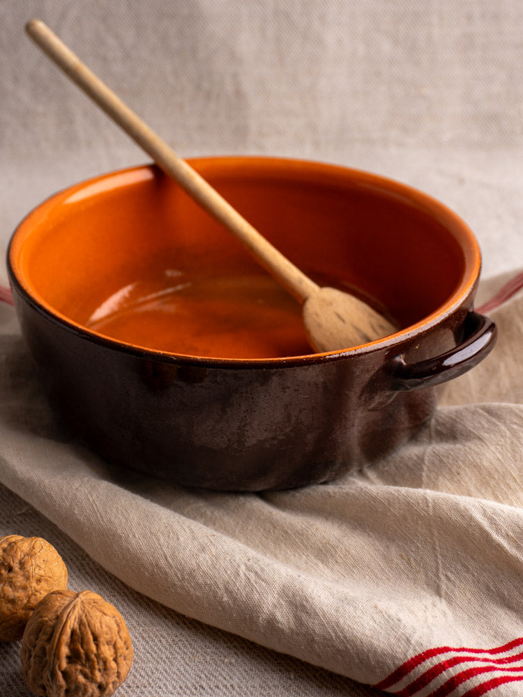Antique French earthenware baking dish with brown and orange glaze, wooden spoon, and walnuts on striped beige cloth.