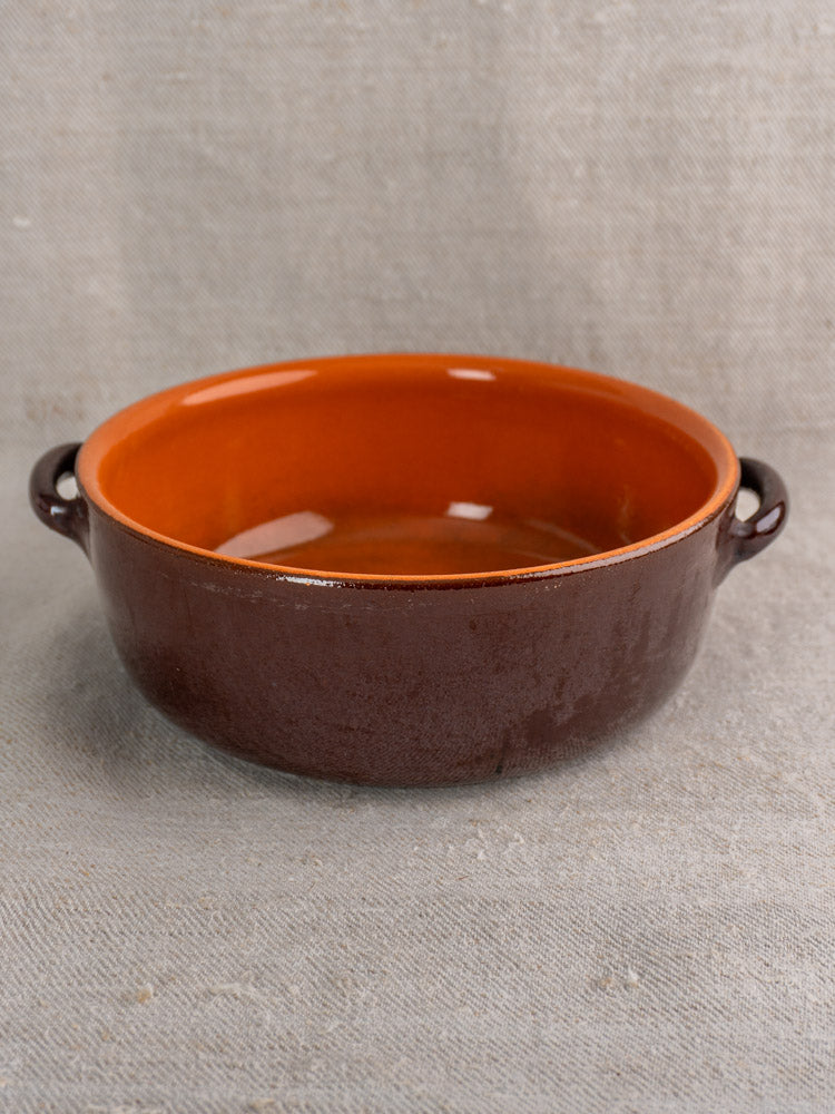 Antique French earthenware baking dish with brown and orange glaze, twin handles, on textured gray fabric.