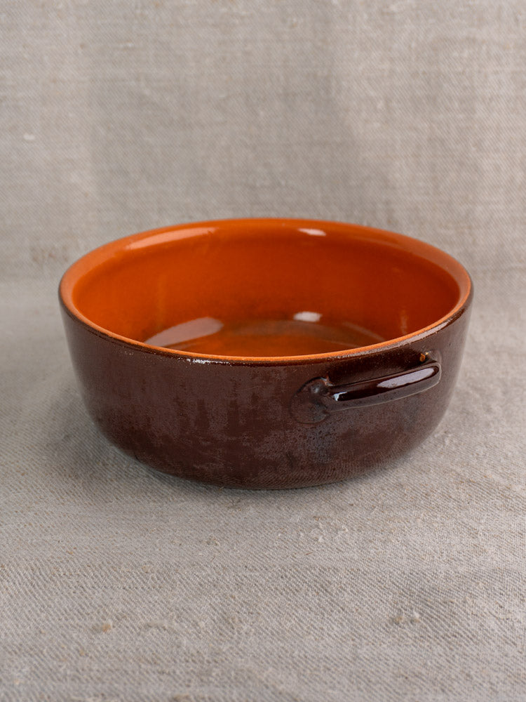 Antique French earthenware baking dish, mid 20th century, with brown and orange glaze, on beige textured fabric.