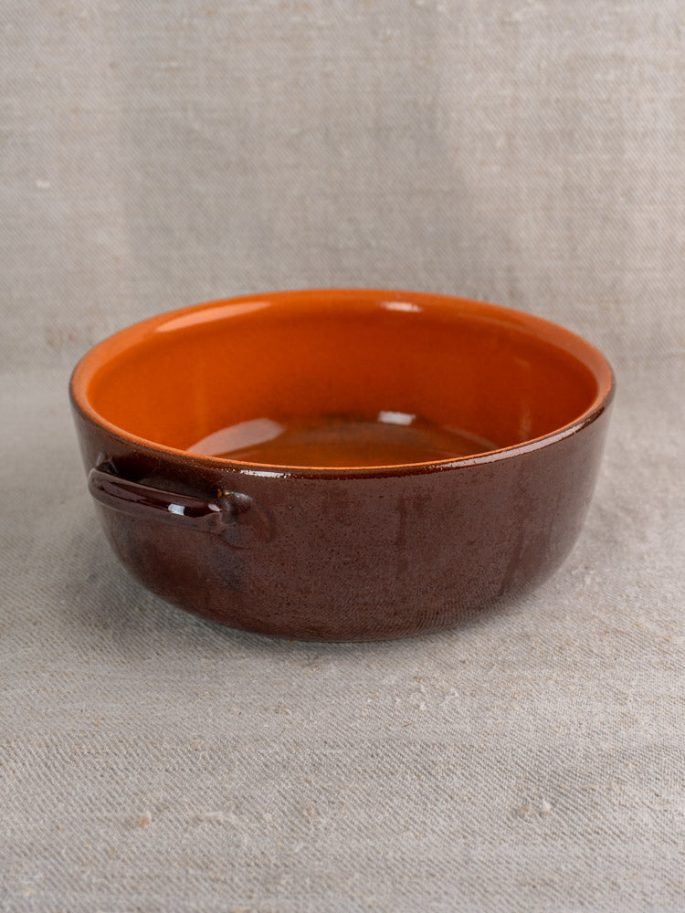 Antique French earthenware baking dish with brown exterior and orange glazed interior on beige textured fabric. Mid 20th century.