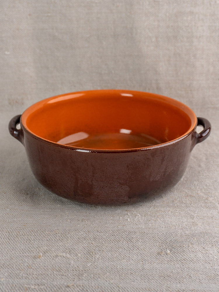 Mid-century French earthenware baking dish with glossy brown glaze, orange interior, and twin handles on beige textured fabric.