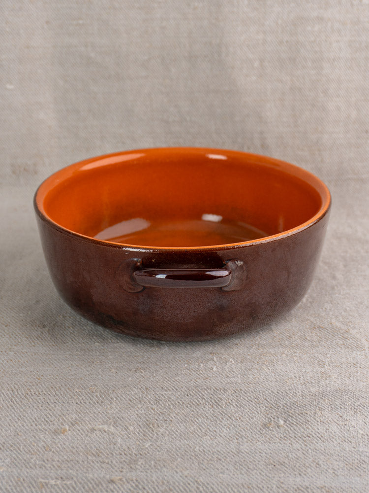 Antique French earthenware baking dish, round with brown-orange glaze, side handles, on textured beige fabric.