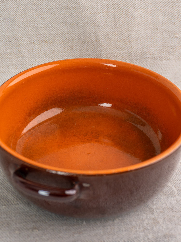 Antique French earthenware baking dish with brown and orange glaze and side handle on light grey textured fabric.