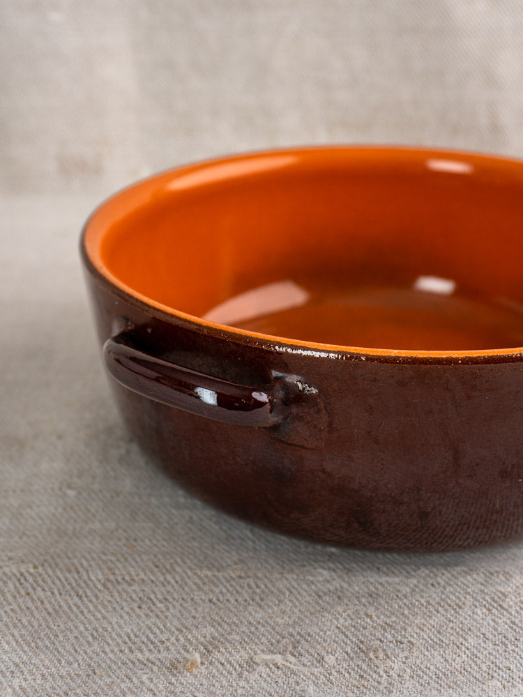 Antique French earthenware baking dish, mid-20th century, with glossy brown-orange glaze and side handle on neutral fabric.