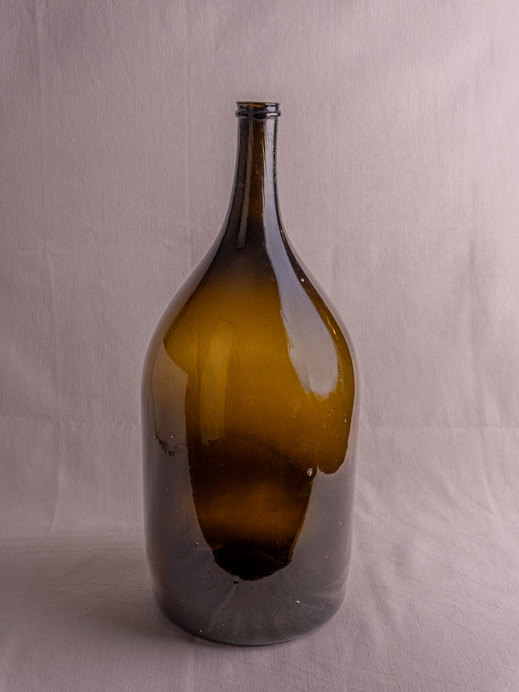 Antique French hand-blown olive green glass wine bottle with long neck on light gray fabric.