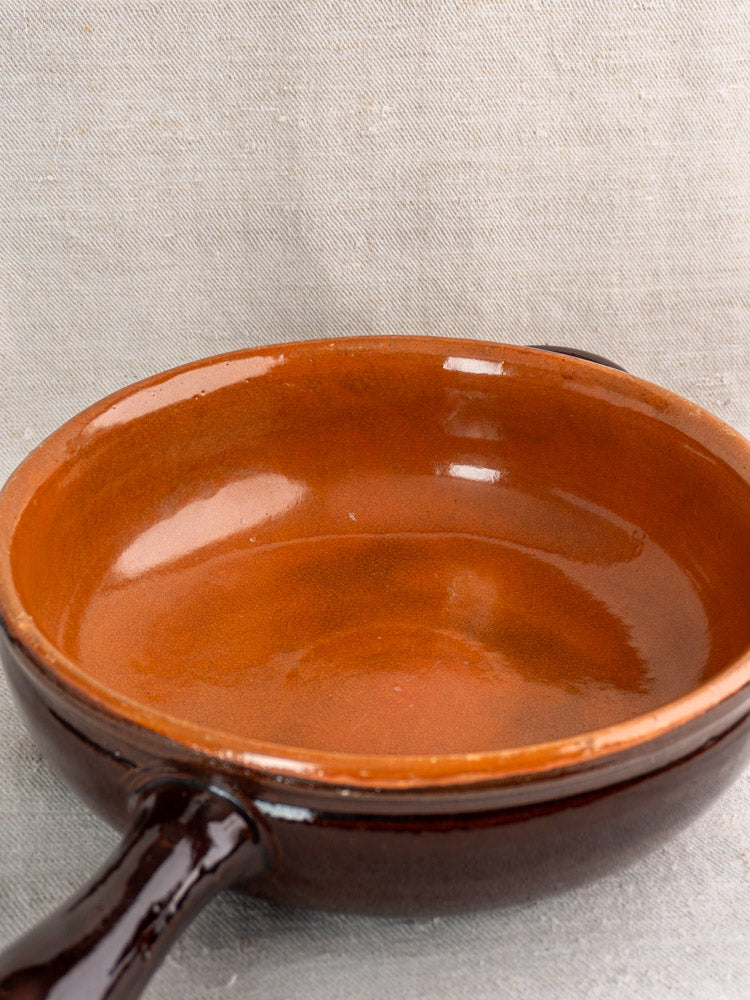 Antique French earthenware poêlon with brown glaze and caramel interior on light gray textured fabric. Glossy, thick-rimmed finish.