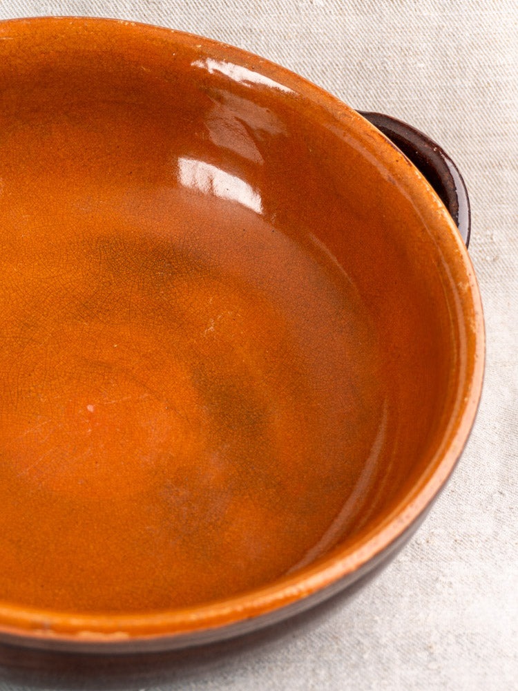 Antique French earthenware poêlon, mid-20th century ceramic dish with brown glaze and small handles on light textured surface.