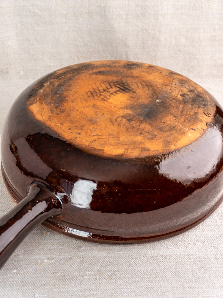 Antique French earthenware poêlon with brown glaze and caramel interior, underside worn, on neutral fabric background.