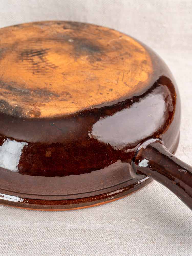 Antique French earthenware poêlon bottom with brown glaze and scorch marks, handle visible right, on light fabric.