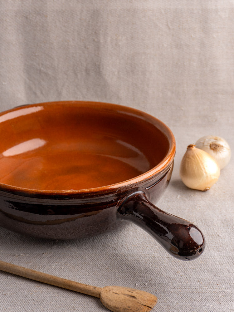 Antique French earthenware poêlon with brown glaze and caramel interior, wooden spatula, and two onions on gray fabric.