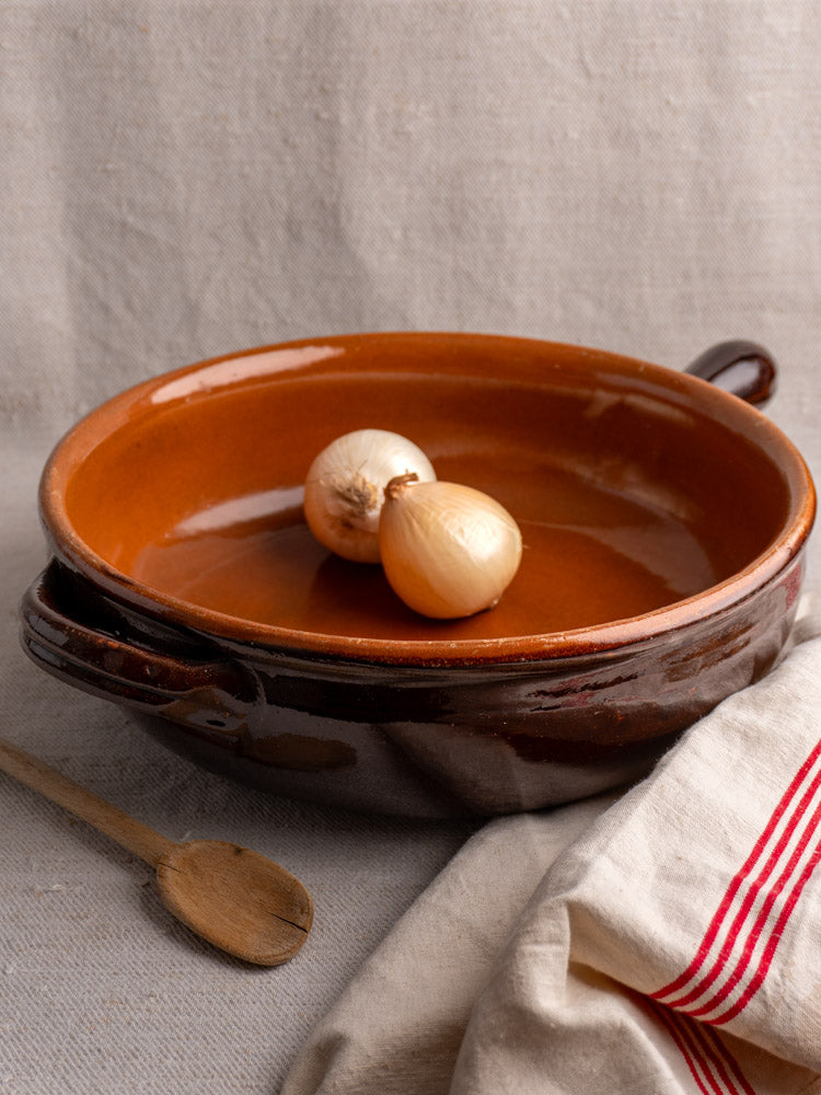 Antique French earthenware poêlon with brown glaze, caramel interior, and yellow onions on light fabric with striped cloth.