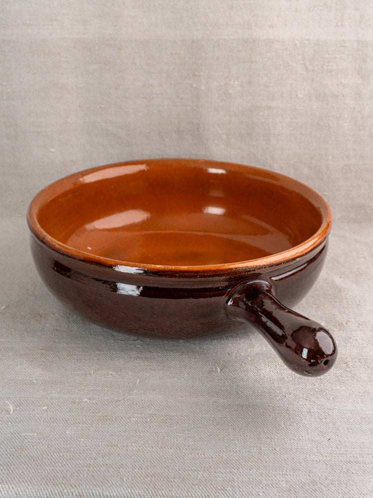 Antique French earthenware poêlon with brown glaze, single handle, caramel interior on textured beige fabric.