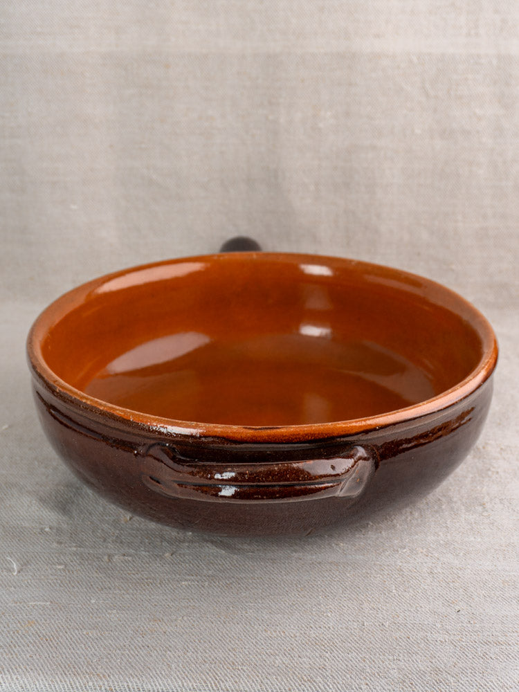 Mid-20th century French earthenware poêlon with brown glaze, caramel interior, and side handle on neutral fabric.