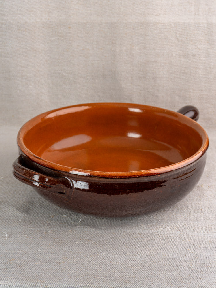 Antique French earthenware poêlon, round and shallow with brown glaze, caramel interior, on neutral fabric. Mid-20th century.