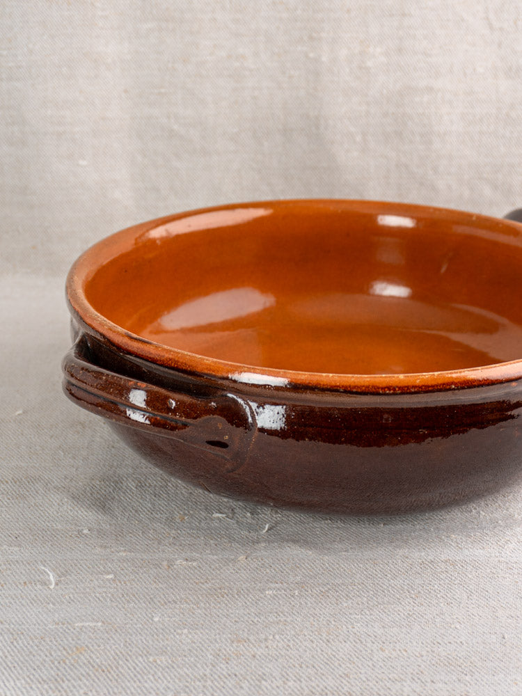 Antique French earthenware poêlon, mid-20th century, brown glaze with caramel interior, on light beige fabric background.