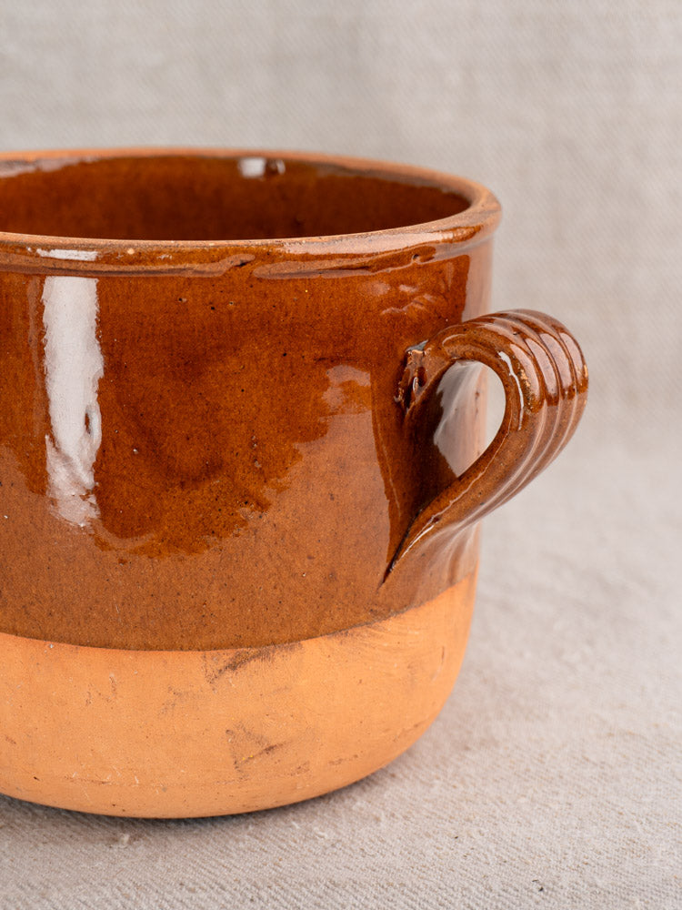 Antique French Digoin confit pot in brown-glazed earthenware with handle and unglazed base on neutral fabric background.