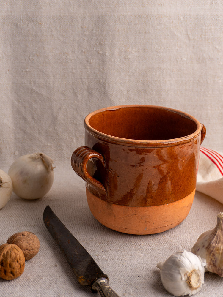 Mid-century French Digoin glazed earthenware confit pot on beige cloth with garlic, walnuts, knife, and red-striped towel.