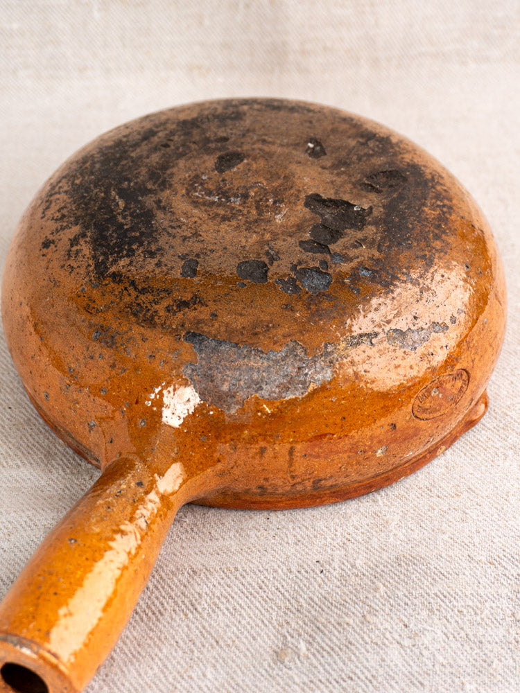 Antique French Vallauris poêlon in glazed earthenware with orange patina and scorch marks, upside down on beige fabric.