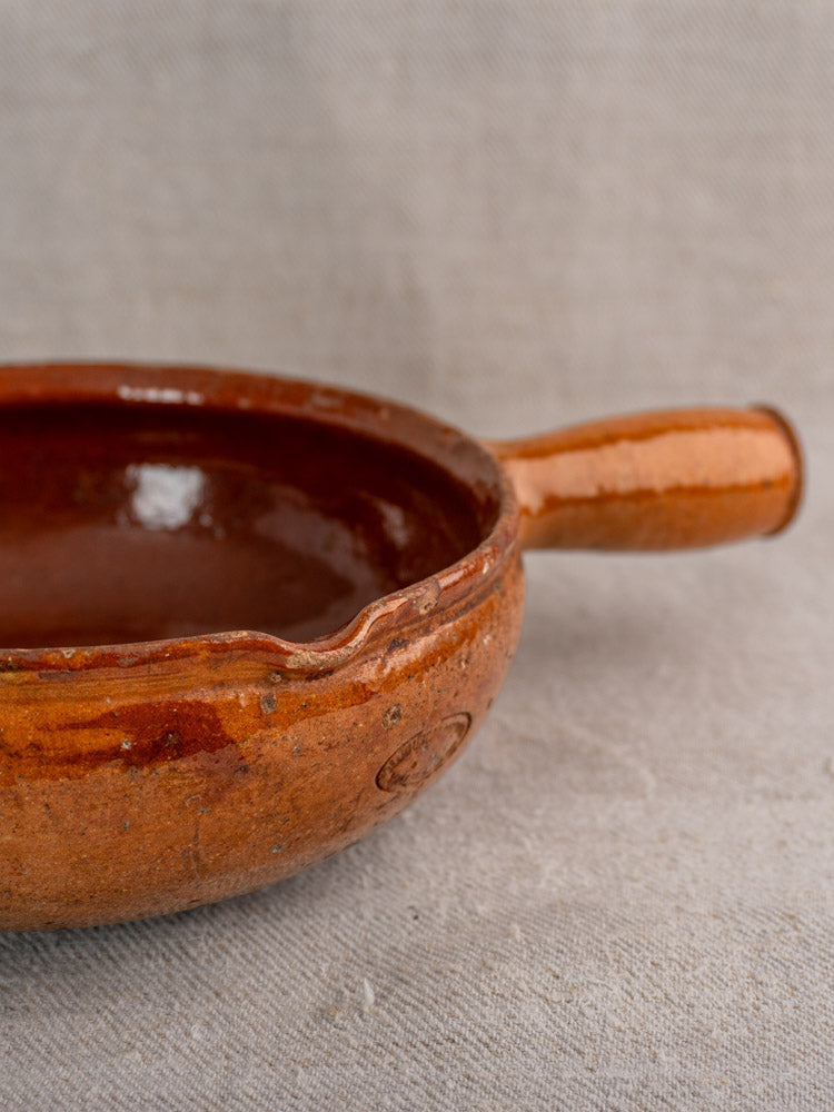 Antique French Vallauris glazed earthenware poêlon with rustic brown finish and handle, slightly chipped rim, on beige fabric.