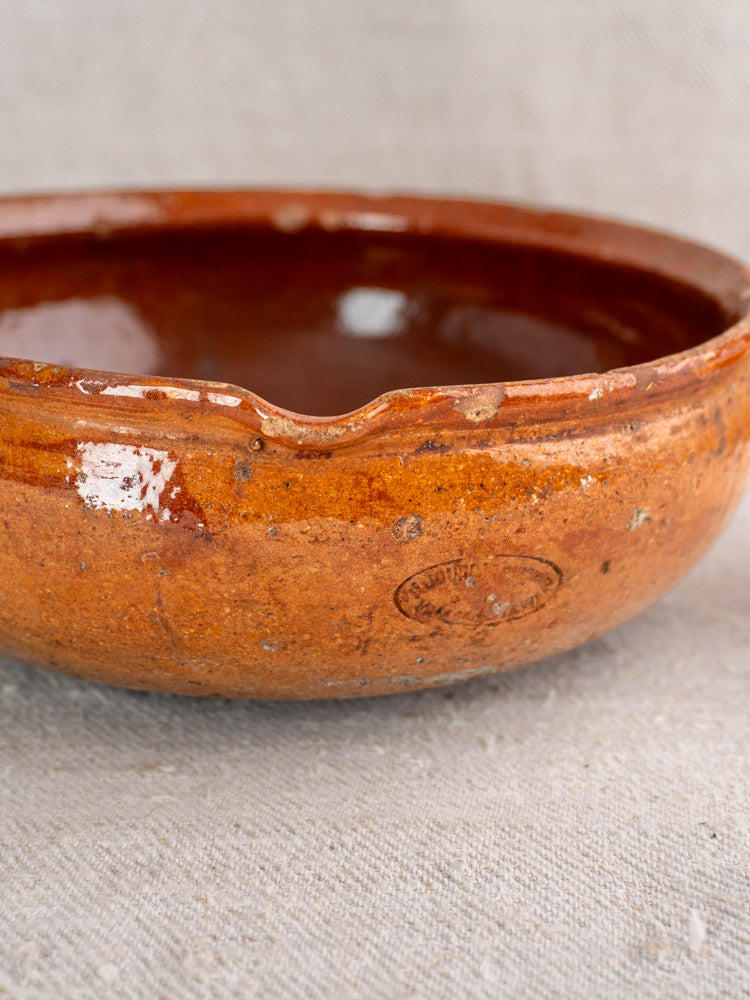 Antique French Vallauris glazed earthenware poêlon with spout and maker’s mark on textured beige fabric.