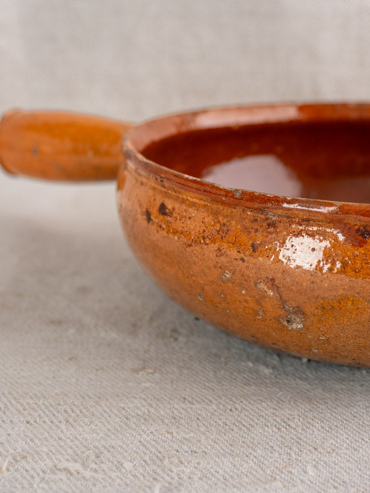 Antique French Vallauris glazed earthenware poêlon with handle, rustic brown glaze, on light textured fabric background.