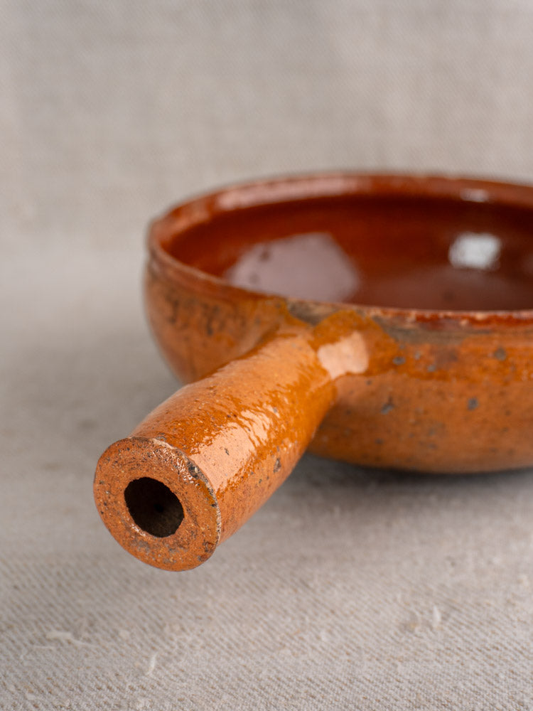 Antique French Vallauris glazed earthenware poêlon, orange-brown ceramic bowl with hollow handle on textured fabric background.