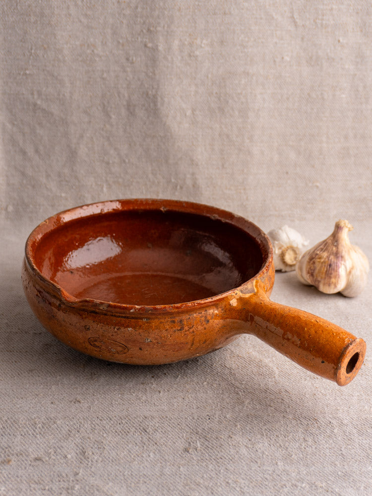 Antique Vallauris French poêlon, glazed earthenware, displayed on beige textured surface with two garlic heads.
