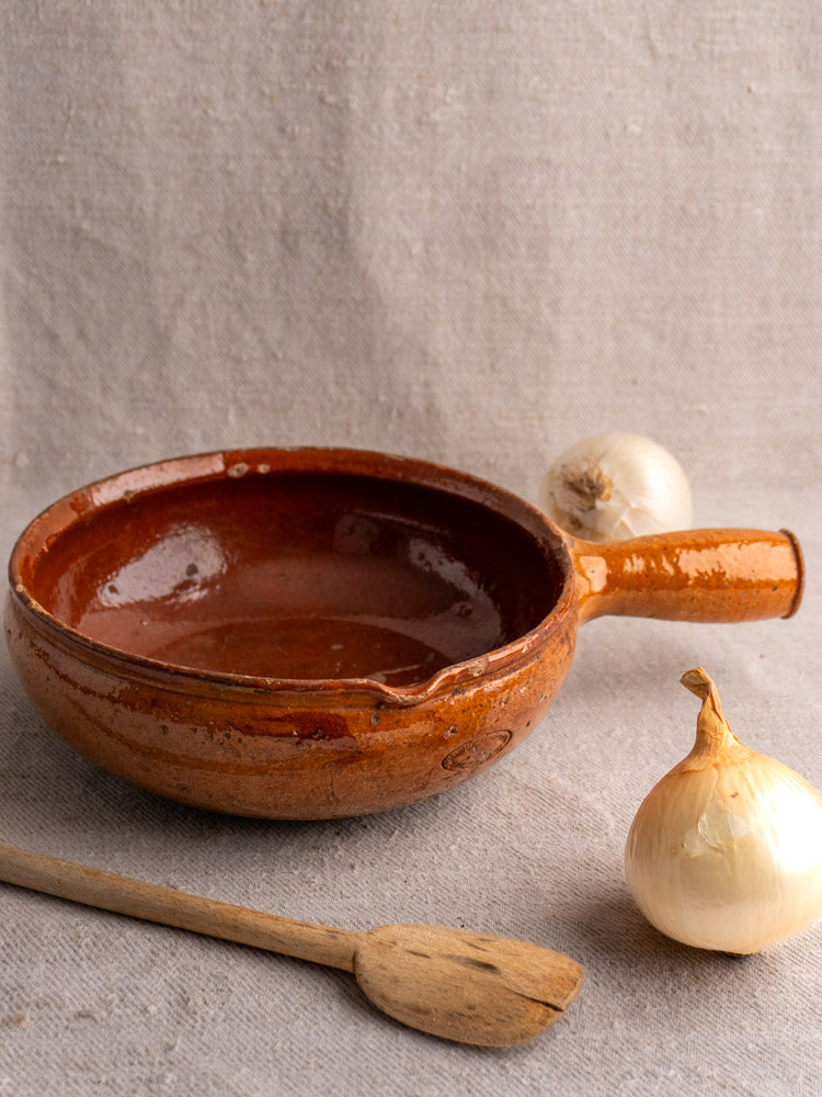 Antique French Vallauris glazed earthenware poêlon with handle on textured fabric, wooden spatula and yellow onions nearby.