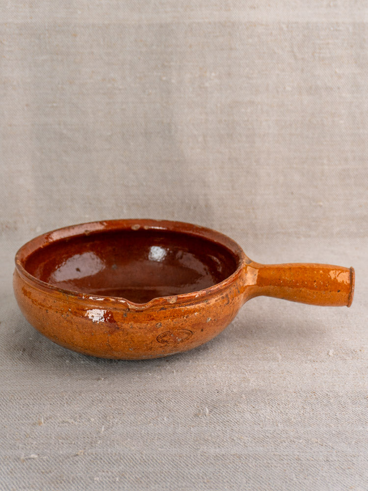 Antique French Vallauris Poêlon, round brown glazed earthenware with handle, glossy finish, on beige textured fabric.